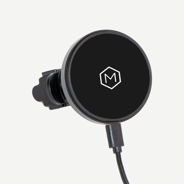 Image of a Mighty Mount MagSafe Wireless Charger attached to a car vent. The charger, designed to hold a smartphone, is circular and sleek, with a visible MagSafe logo at the center. The car's dashboard and a portion of the steering wheel are slightly visible in the background, suggesting the charger's placement for easy access and visibility while driving.